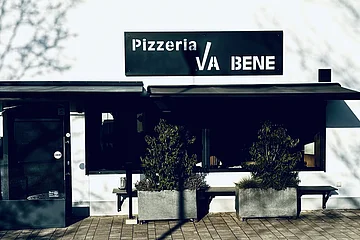 Außenansicht einer Pizzeria mit schwarzem Schild „Pizzeria VA BENE“, eine Eingansbereich aus Glas, verdunkeltem großen Fenster mit Rollläden und zwei großen Pflanzkübeln davor. Vor der Pizzeria ein gepflasterter Gehweg.