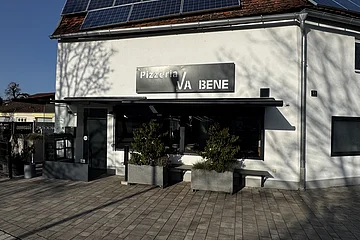 Außenansicht einer Pizzeria mit schwarzem Schild „Pizzeria VA BENE“, eine Eingangsbereich aus Glas, verdunkeltem großen Fenster mit Rollläden und zwei großen Pflanzkübeln davor. Vor der Pizzeria ein gepflasterter Gehweg. Auf dem Dach des  Gebäudes viele Solarpanele.
