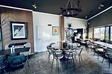 Moderner Innenraum einer Pizzeria mit Tischen, Stühlen, Wandbildern und einer Getränkekarte an der Wand. Größtenteils in grau-weiß eingerichtet mit Holzoptik-Fassaden an der Wand.