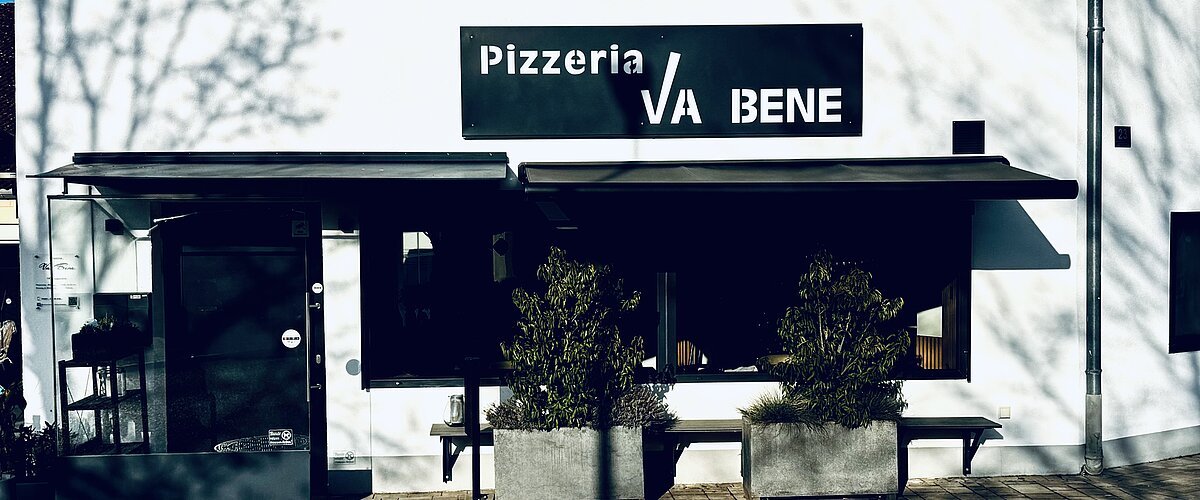 Außenansicht einer Pizzeria mit schwarzem Schild „Pizzeria VA BENE“, eine Eingansbereich aus Glas, verdunkeltem großen Fenster mit Rollläden und zwei großen Pflanzkübeln davor. Vor der Pizzeria ein gepflasterter Gehweg.