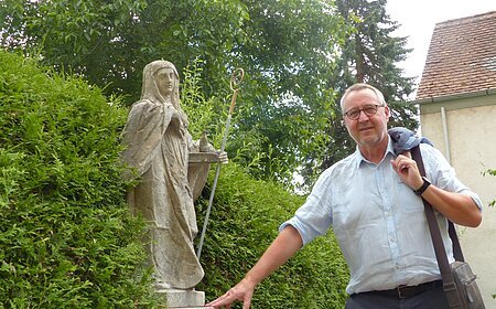 Eine Person mit Jacke über der Schulter steht neben einer Statue in einem Garten, umgeben von Büschen und einem Gebäude im Hintergrund.