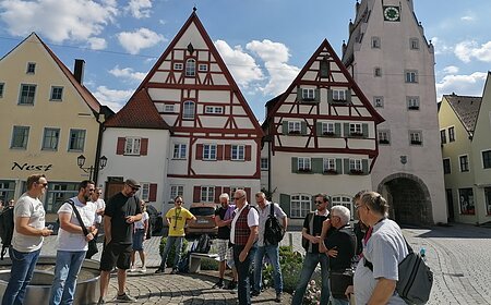 Gruppe von Menschen steht auf einem gepflasterten Platz, der Monheimer Innenstadt, vor Fachwerkhäusern und einem Turm mit Uhr. Auf einem gelben Haus davon steht der Schriftzug Nest.