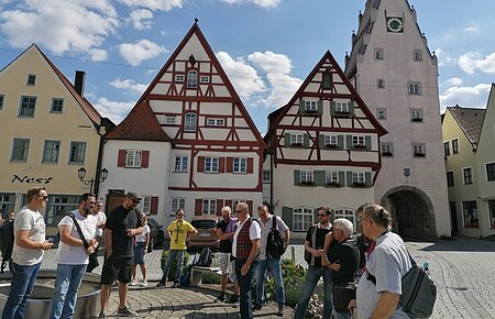 Gruppe von Menschen steht auf einem gepflasterten Platz, der Monheimer Innenstadt, vor Fachwerkhäusern und einem Turm mit Uhr. Auf einem gelben Haus davon steht der Schriftzug Nest.