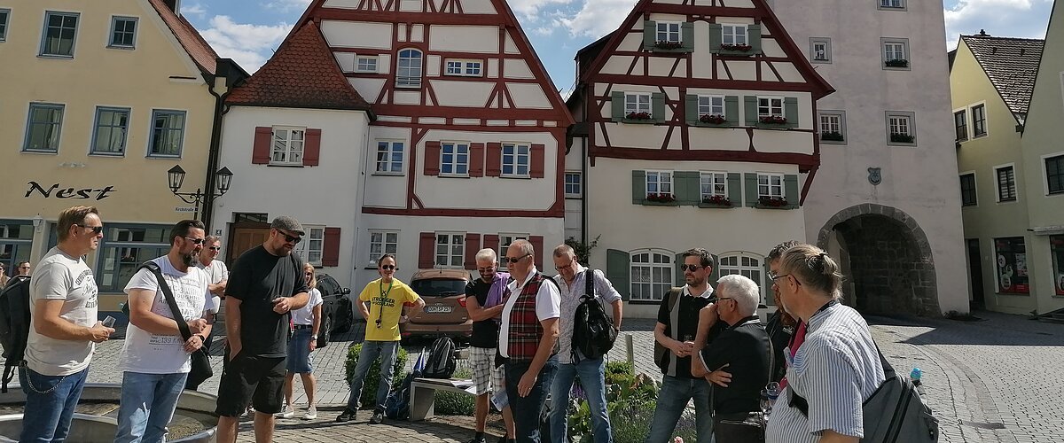 Gruppe von Menschen steht auf einem gepflasterten Platz, der Monheimer Innenstadt, vor Fachwerkhäusern und einem Turm mit Uhr. Auf einem gelben Haus davon steht der Schriftzug Nest.