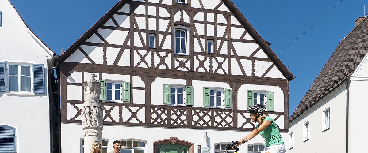 Der Marktplatz in Monheim: ein Fachwerkhaus zwischen zwei Gebäuden, ein Fahrradfahrer im Vordergrund fährt auf dem gepflastertem Platz, zwei Personen sitzen auf dem Rand eines Brunnens vor dem Fachwerkhaus. Auf dem linken Gebäude steht die Aufschrift "Stadtapotheke", auf dem rechten Gebäude die Aufschrift "Sparkasse".