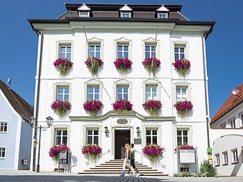 Das Rathaus in Monheim: Ein weißes dreistöckiges Gebäude mit mehreren Fenstern, an denen Blumenkästen mit pinken Blumen hängen. Zwei Personen gehen davor vorbei. Links und rechts davon jeweils ein weiteres Haus.