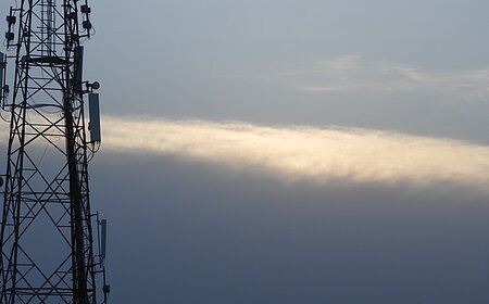 Mobilfunk-Sendemast vor bewölktem Himmel mit einer schmalen, hellen Wolke im Hintergrund.