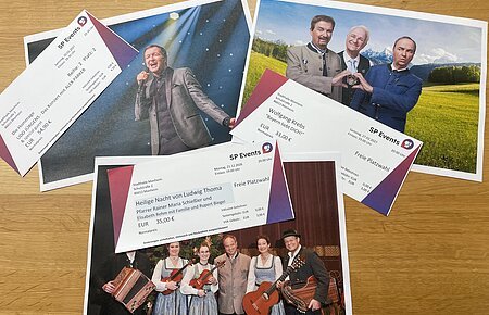 Drei Konzertkarten von SP Events liegen auf Fotos von Musikern in traditioneller und moderner Kleidung auf einem Holztisch. Kartenvorverkauf für die nächsten Veranstaltungen in Monheim