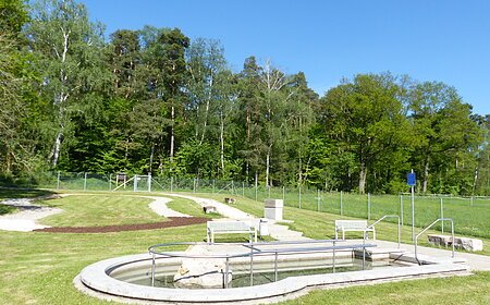 Rundes Kneippbecken in Monheim mit einem Geländer und einem Barfußpfad auf einer Wiese, umgeben von einer großen Grünfläche und Bäumen und Sitzmöglichkeiten. Ein Zaun im Hintergrund.