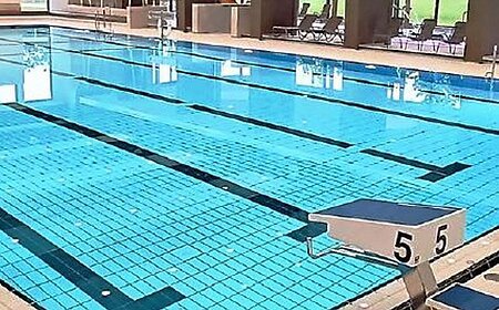 Titelseite des Flyers "BÄDER MONHEIM" mit dem gleichnamigen Titel in weiß/blau auf gelbem Hintergrund oben im Bild. Darunter die Innenaufnahme eines Schwimmbads mit einem gefülltem Schwimmbecken, daneben der Startblock Nummer 5, ein Wasserspielball und Sitzbereich im Hintergrund. Unten links im Eck die Angabe der Webseite "www.monheim-bayern.de".