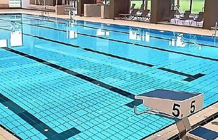 Titelseite des Flyers "BÄDER MONHEIM" mit dem gleichnamigen Titel in weiß/blau auf gelbem Hintergrund oben im Bild. Darunter die Innenaufnahme eines Schwimmbads mit einem gefülltem Schwimmbecken, daneben der Startblock Nummer 5, ein Wasserspielball und Sitzbereich im Hintergrund. Unten links im Eck die Angabe der Webseite "www.monheim-bayern.de".