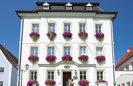 Das Rathaus der Drei-Stämme-Stadt Monheim Das Rathaus in Monheim: Ein weißes dreistöckiges Gebäude mit mehreren Fenstern, an denen Blumenkästen mit pinken Blumen hängen. Zwei Personen gehen davor vorbei. Links und rechts davon jeweils ein weiteres Haus.