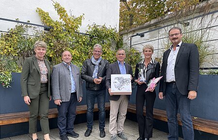 Dienstjubiläum Elisabeth Besold Sechs Personen stehen nebeneinander vor einer hohen Beet in anthrazit mit Pflanzen, darunter Bänke entlang des Beetes. Zwei Personen halten Geschenke in den Händen.