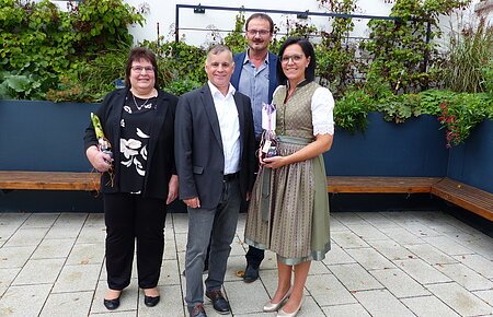 Dienstjubiläum Sandra Steidle und Erika Roßkopf Vier Personen stehen auf gepflastertem Hof vor einem schwarzen Hochbeet mit grünen Pflanzen und Bänken entlang des Beetes, zwei Personen halten Geschenke.
