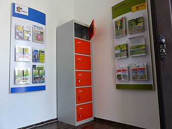 Ein orange-grauer Indoor-Ladeschrank für eBike-Akkus mit insgesamt sechs Fächern und einem offenen Fach steht zwischen zwei Wanddisplays mit Broschüren in dem Vorraum der Touristinformation Monheim.