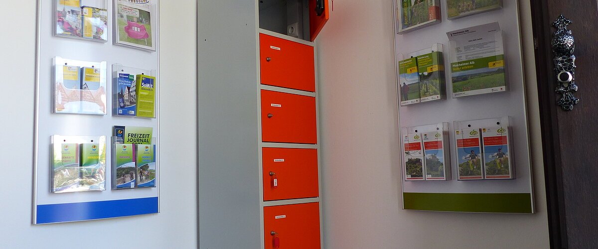 Ein orange-grauer Indoor-Ladeschrank für eBike-Akkus mit insgesamt sechs Fächern und einem offenen Fach steht zwischen zwei Wanddisplays mit Broschüren in dem Vorraum der Touristinformation Monheim.