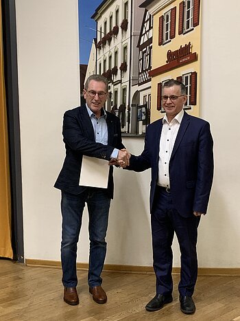 Zwei Männer in Anzügen schütteln sich die Hand vor einer Wand mit einem Bild von Fachwerkhäusern. Der Mann links hält eine Urkunde in der anderen Hand.
