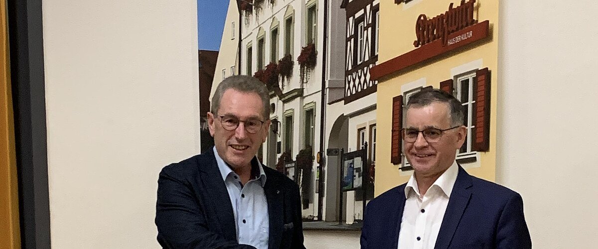 Zwei Männer in Anzügen schütteln sich die Hand vor einer Wand mit einem Bild von Fachwerkhäusern. Der Mann links hält eine Urkunde in der anderen Hand.