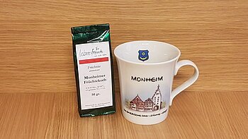 Geschenkidee der Stadt Monheim: eine weiße Tasse mit Monheim-Motiv und der Aufschrift "Monheim - Die Drei-Stämme-Stadt" rechts im Bild. Links davon eine Teeverpackung mit der Mischung und dem Text „Monheimer Früchtekorb“ stehen vor einer Holzoberfläche.