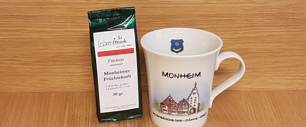 Geschenkidee der Stadt Monheim: eine weiße Tasse mit Monheim-Motiv und der Aufschrift "Monheim - Die Drei-Stämme-Stadt" rechts im Bild. Links davon eine Teeverpackung mit der Mischung und dem Text „Monheimer Früchtekorb“ stehen vor einer Holzoberfläche.