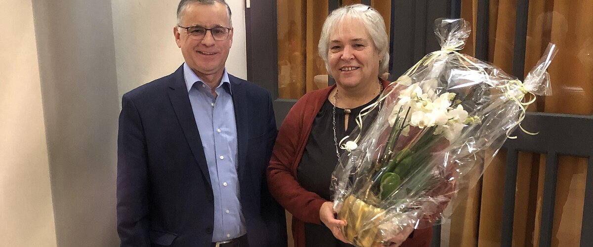 Zwei Personen stehen nebeneinander in einem Raum mit Fliesenbelag und einer schwarzen Tür mit orangem Vorhang im Hintergrund. Die Frau hält einen großen weißen verpackten Blumenstock.