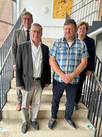 Vier Personen stehen blickend in die Kamera auf einer Treppe in einem Gebäude mit schwarzem Geländer und großem Fenster. An der Wand hängt ein gelbes Gemälde.