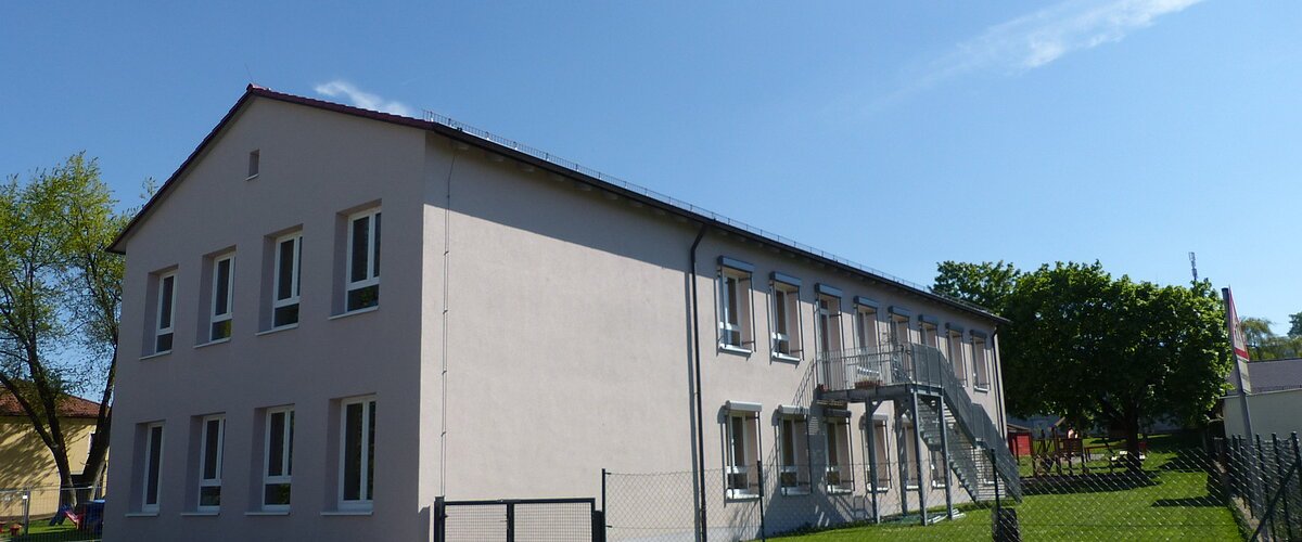 Städtischer Kindergarten in Monheim: ein zweistöckiges Gebäude mit weißer Fassade, Fenstern und Außentreppe, umgeben von einem Zaun und Bäumen. Rechts um das Gebäude ist ein Garten zu sehen. Vorderhalb des Gebäudes verläuft eine Straße.