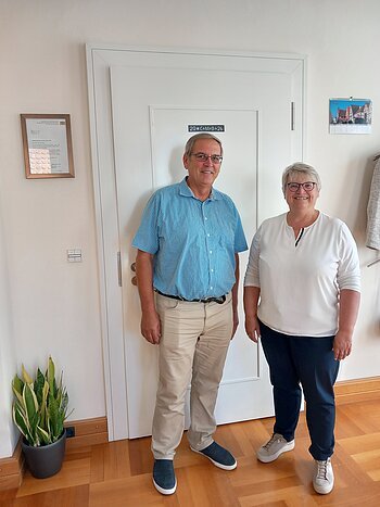 Mann und Frau stehen vor einer weißen Tür in einem Raum mit Holzboden und Pflanzen. Erste Bürgermeisterin Anita Ferber bei der Vereidigung des neuen Feldgeschworenen für den Stadtteil Flotzheim Hermann Färber