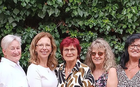 Neun Frauen stehen eng nebeneinander vor einer grünen Hecke, im Vordergrund Tische mit Stühlen und Besteck. Ehrenamtliches Helferteam Soziales Miteinander - 10 Jahre Fundgrube Monheim