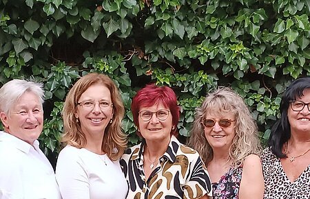 Soziales Miteinander - 10 Jahre Fundgrube Monheim Neun Frauen stehen eng nebeneinander vor einer grünen Hecke, im Vordergrund Tische mit Stühlen und Besteck. Ehrenamtliches Helferteam Soziales Miteinander - 10 Jahre Fundgrube Monheim