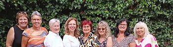 Neun Frauen stehen eng nebeneinander vor einer grünen Hecke, im Vordergrund Tische mit Stühlen und Besteck. Ehrenamtliches Helferteam Soziales Miteinander - 10 Jahre Fundgrube Monheim