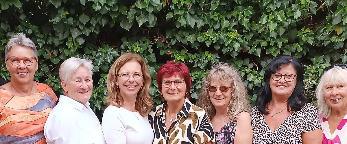 Neun Frauen stehen eng nebeneinander vor einer grünen Hecke, im Vordergrund Tische mit Stühlen und Besteck. Ehrenamtliches Helferteam Soziales Miteinander - 10 Jahre Fundgrube Monheim