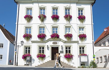Das Rathaus der Drei-Stämme-Stadt Monheim Das Rathaus in Monheim: Ein weißes dreistöckiges Gebäude mit mehreren Fenstern, an denen Blumenkästen mit pinken Blumen hängen. Zwei Personen gehen davor vorbei. Links und rechts davon jeweils ein weiteres Haus.