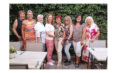 Neun Frauen stehen eng nebeneinander vor einer grünen Hecke, im Vordergrund Tische mit Stühlen und Besteck. Ehrenamtliches Helferteam Soziales Miteinander - 10 Jahre Fundgrube Monheim