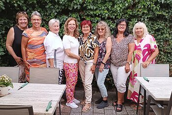 Neun Frauen stehen eng nebeneinander vor einer grünen Hecke, im Vordergrund Tische mit Stühlen und Besteck. Ehrenamtliches Helferteam Soziales Miteinander - 10 Jahre Fundgrube Monheim