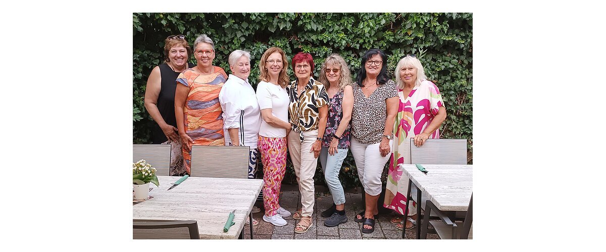 Neun Frauen stehen eng nebeneinander vor einer grünen Hecke, im Vordergrund Tische mit Stühlen und Besteck. Ehrenamtliches Helferteam Soziales Miteinander - 10 Jahre Fundgrube Monheim
