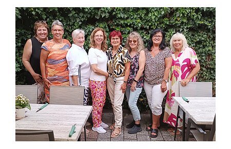Soziales Miteinander - 10 Jahre Fundgrube Monheim Neun Frauen stehen eng nebeneinander vor einer grünen Hecke, im Vordergrund Tische mit Stühlen und Besteck. Ehrenamtliches Helferteam Soziales Miteinander - 10 Jahre Fundgrube Monheim