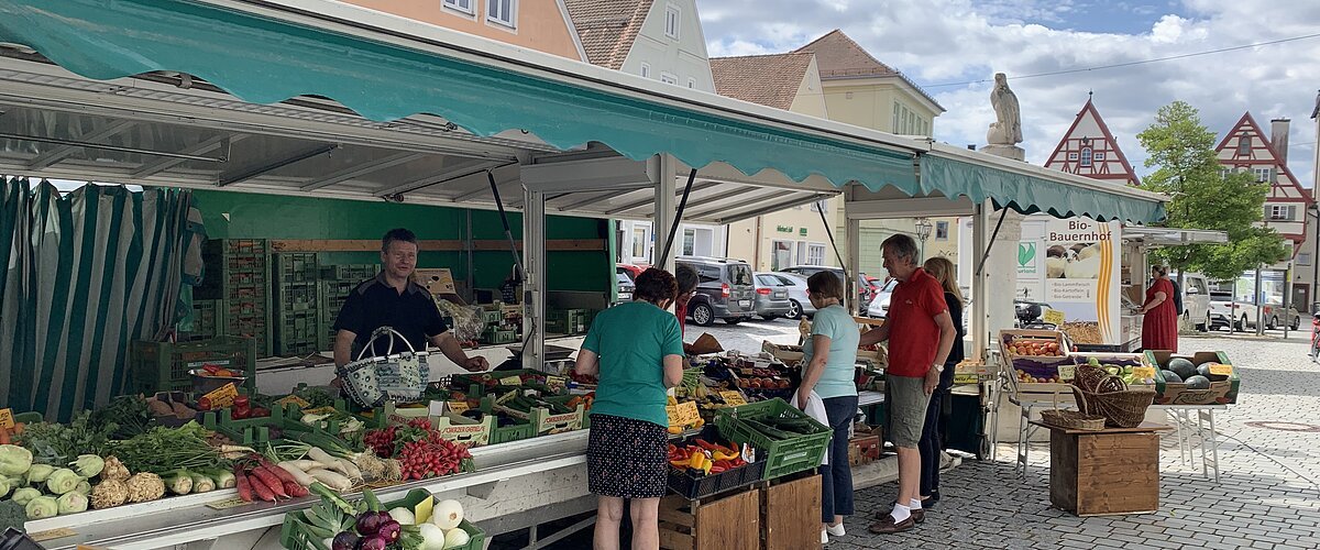 Marktstand des Wochenmarkts in Monheim mit Obst und Gemüse in der Auslage auf dem gepflastertem Marktplatz. Mehrere Personen kaufen dort ein. Fachwerkhäuser im Hintergrund und bewölkter Himmel.