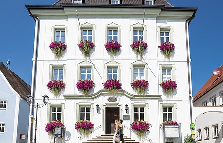 Das Rathaus der Drei-Stämme-Stadt Monheim_ Das Rathaus in Monheim: Ein weißes dreistöckiges Gebäude mit mehreren Fenstern, an denen Blumenkästen mit pinken Blumen hängen. Zwei Personen gehen davor vorbei. Links und rechts davon jeweils ein weiteres Haus.