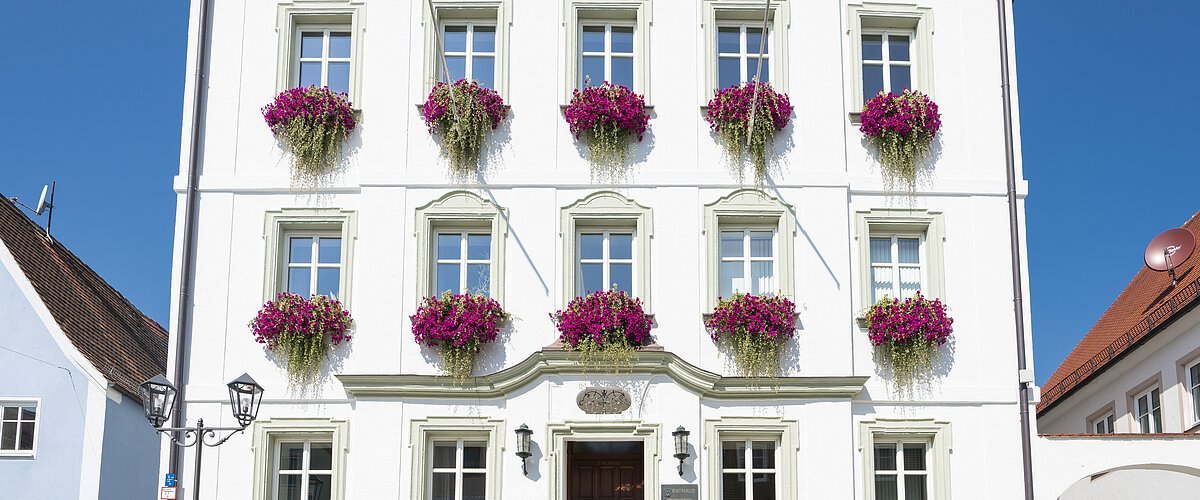 Das Rathaus in Monheim: Ein weißes dreistöckiges Gebäude mit mehreren Fenstern, an denen Blumenkästen mit pinken Blumen hängen. Zwei Personen gehen davor vorbei. Links und rechts davon jeweils ein weiteres Haus.