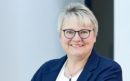 Porträt einer lächelnden Frau mit kurzen grauen Haaren, Brille, weißem Hemd und blauem Blazer, die an einem Geländer lehnt.
1. Bürgermeisterin der Stadt Monheim