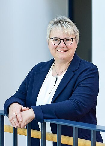 Porträt einer lächelnden Frau mit kurzen grauen Haaren, Brille, weißem Hemd und blauem Blazer, die an einem Geländer lehnt.
1. Bürgermeisterin der Stadt Monheim