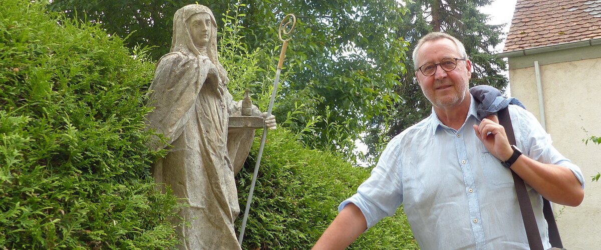 Eine Person mit Jacke über der Schulter steht neben einer Statue in einem Garten, umgeben von Büschen und einem Gebäude im Hintergrund.