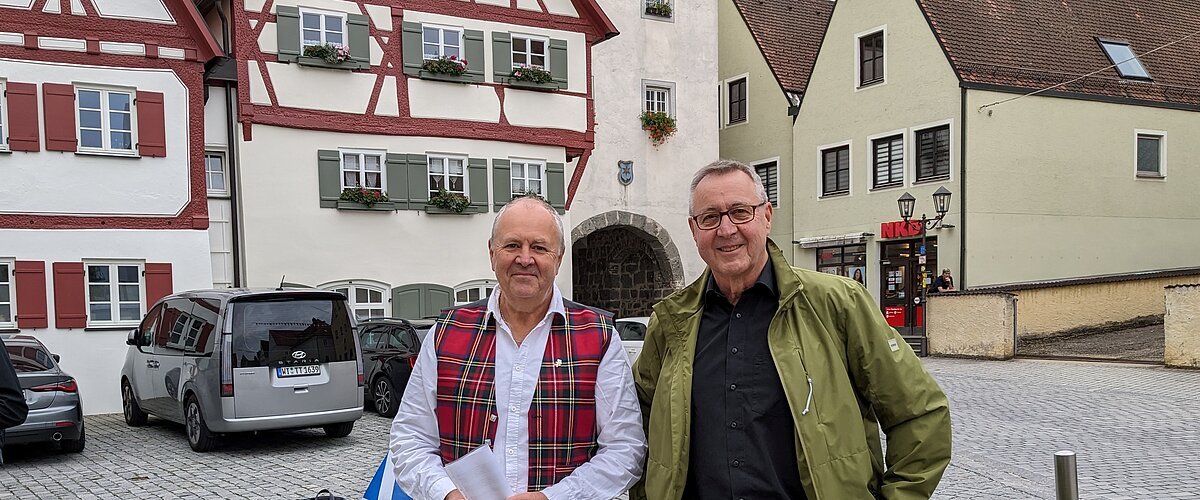 Zwei Männer stehen vor Fachwerkhäusern auf gepflastertem Platz und einer Bank mit Rucksack und Flaschen im Hintergrund. Die Bank ist umgeben von Pflanzen. Eine schottische Fahne - in weiß-blau - liegt auf der Sitzbank.