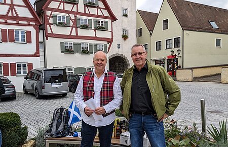 Monheimer Whisky-Tour Stadtführer Rainer Keßler & Whisky-Liebhaber H.P. Schenk Zwei Männer stehen vor Fachwerkhäusern auf gepflastertem Platz und einer Bank mit Rucksack und Flaschen im Hintergrund. Die Bank ist umgeben von Pflanzen. Eine schottische Fahne - in weiß-blau - liegt auf der Sitzbank.