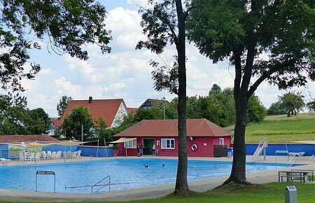 Freibad Monheim - Schwimmbecken & Kiosk Ein Freibad mit Schwimmbecken, einer Rutsche und Sitzmöglichkeiten, umgeben von Bäumen und Wiesen. Im Hintergrund steht ein rotes Gebäude mit Fenstern, ein Kiosk.