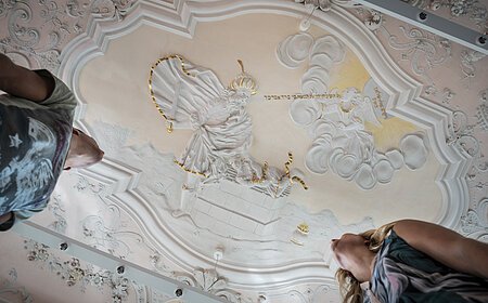 Stuckdecke im Monheimer Rathaus mit barocker farbiger Stuckreliefdarstellung und zwei Personen, die von unten darauf schauen.