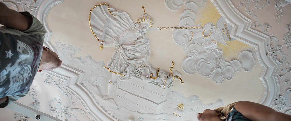Stuckdecke im Monheimer Rathaus mit barocker farbiger Stuckreliefdarstellung und zwei Personen, die von unten darauf schauen.