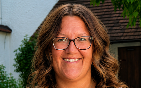 Porträt einer lächelnden Frau mit Brille, braunen lockigen Haaren und floralem Oberteil vor Hausfassade und Grün. Erste Jugendbeauftragte der Stadt Monheim: Kathrin Böswald