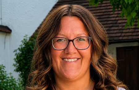 Jugendbeauftragte Kathrin Böswald Porträt einer lächelnden Frau mit Brille, braunen lockigen Haaren und floralem Oberteil vor Hausfassade und Grün. Erste Jugendbeauftragte der Stadt Monheim: Kathrin Böswald